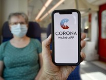 Hand hält Smartphone mit Corona Warn-App vor ältere Frau mit Mundschutzmaske sitzt in S-Bahn, Zug, Corona-Krise, Stuttg
